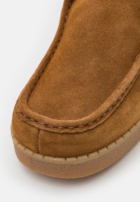 Pantofole in suede marrone con punta arrotondata, cuciture visibili e suola in gomma texture. Presenta una finitura morbida e un design minimale.