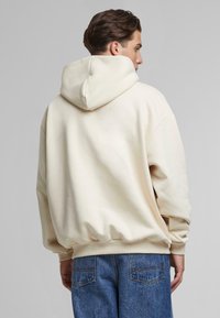 Hoodie oversize color crema con cappuccio, polsini e orlo a costine. Texture morbida, schiena senza cuciture, abbinato a jeans in denim blu scuro.