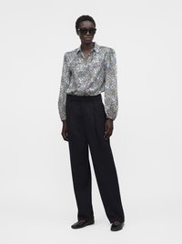 Femme portant de grandes lunettes de soleil noires, un chemisier à manches longues à fleurs, un pantalon noir taille haute à jambes larges, et des ballerines noires, debout devant un fond blanc.