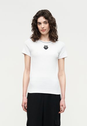 Γυναίκα με σγουρά μαλλιά που φοράει λευκό πουκάμισο με κοντά μανίκια και μαύρο έμβλημα, καθώς και μαύρο παντελόνι, μπροστά σε ανοιχτό φόντο.