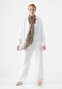 Witte linnen shirt over een witte tanktop, gecombineerd met losse witte broeken. Bruine luipaardprint sjaal om de nek gedrapeerd, minimalistische sandalen.