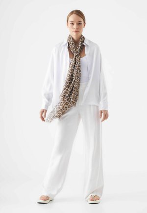 Chemise en lin blanche sur un débardeur blanc, associée à un pantalon large blanc. Écharpe à imprimé léopard marron drapée autour du cou, sandales minimalistes.