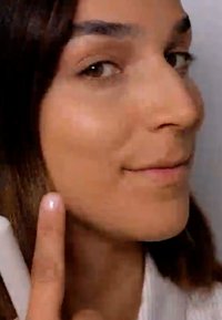 Gros plan du visage d'une personne au teint clair, avec une texture lisse, de longs cheveux bruns et un doigt pointant vers la joue, sans accessoires.