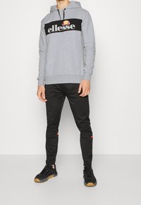 Sweat à capuche gris avec bande noire et logo « ellesse », associé à un pantalon noir avec des accents rouges, et des baskets noires à semelles en gomme.