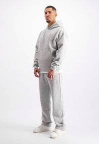 Man staat, draagt een lichtgrijze hoodie en bijpassende sweatpants met witte sneakers, tegen een effen witte achtergrond.