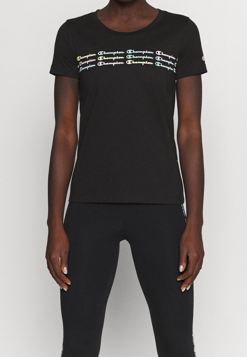 Camiseta negra de manga corta con logos de "Champion" de colores repetidos en el pecho, llevada por una persona de pie con los brazos relajados.