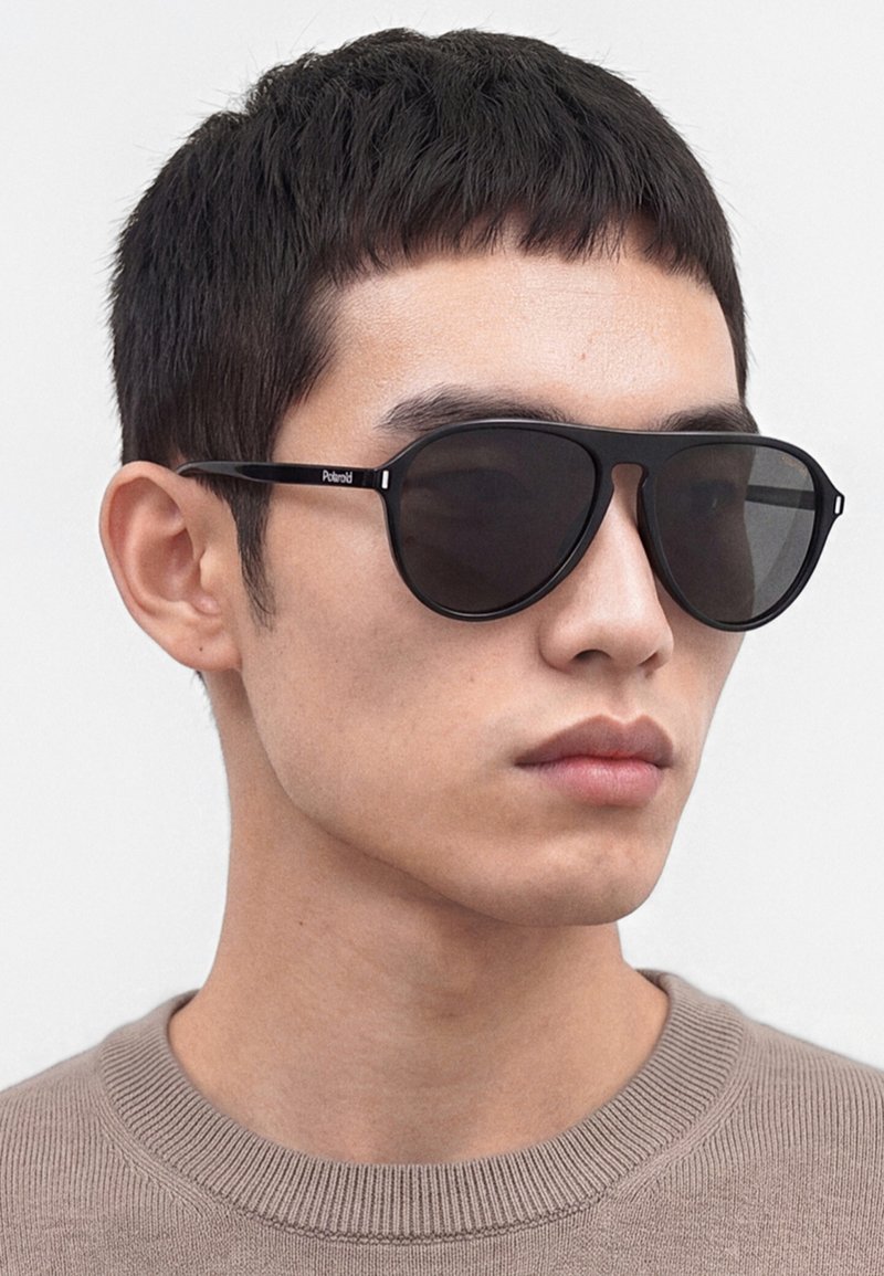 Uomo con capelli corti e neri che indossa occhiali da sole Polaroid neri e un maglione girocollo marrone chiaro, guarda di lato su uno sfondo bianco.