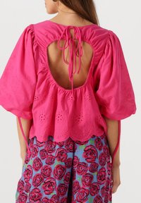 Roze katoenen blouse met pofmouwen, oogjesdetails en een grote uitsnede aan de achterkant afgewerkt met strikjes. Gepaard met een rok met bloemenpatroon.