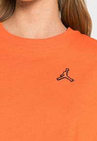 Pomarańczowa bawełniana koszulka z okrągłym dekoltem i krótkimi rękawami, z czarnym haftowanym logo Jumpman na piersi. Gładka tekstura.