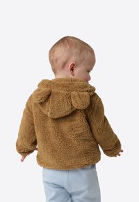 Jachetă maro, pufoasă, cu glugă, realizată din fleece texturat, cu urechi pe glugă, purtată de un copil văzut de la spate. Pantaloni albastru deschis vizibili.