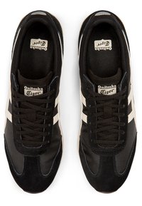 Des chaussures de sport noires avec des rayures blanches, en maille et en daim, présentant un design à lacets et une semelle en caoutchouc texturée avec un logo à l'intérieur.