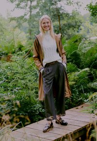 Veste imperméable marron sur un pull en tricot blanc, pantalon gris et chaussures compensées noires. Foliage vert et un chemin en bois en arrière-plan.