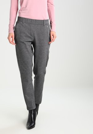 Kvinde iført teksturerede grå tætsiddende bukser, lyserød langærmet top og sorte ankelstøvler, stående mod en ensfarvet lys baggrund.