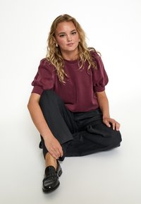 Joven con cabello rubio rizado, vistiendo una blusa burgundy de mangas abullonadas, pantalones de rayas oscuros y mocasines negros, sentada en el suelo.