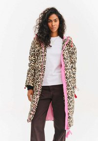 Pružný kabát s leopardím vzorem a růžovým zipem a detaily s tkaničkami, s dlouhými rukávy a délkou až po kolena, nošený přes bílou košili.