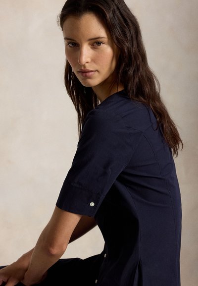 Νεαρή γυναίκα με μακριά σκούρα μαλλιά, ντυμένη με μπλε ναυτικό πουκάμισο με κοντό μανίκι και κουμπιά, καθισμένη και κοιτάζοντας προς την κάμερα, πάνω σε απλό φόντο.