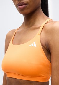 Κοντινό πλάνο ενός ατόμου που φοράει πορτοκαλί αθλητικό σουτιέν με λεπτές τιράντες και λευκό λογότυπο Adidas στο αριστερό μέρος.