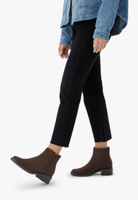 Person som bär bruna ankelstövlar, svarta åtsittande byxor och en blå denimjacka, går på en vit bakgrund.
