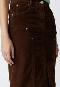 Smeđa kedra suknja s vertikalnim rebrima, srebrnim gumbima i bočnim džepovima. Sadrži visoki prorez za dodatni detalj.