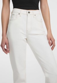 Jeans blancos de cintura alta hechos de un grueso denim de algodón, con bolsillos delanteros, cierre de botón y un detalle de costura contrastante a lo largo de las costuras.