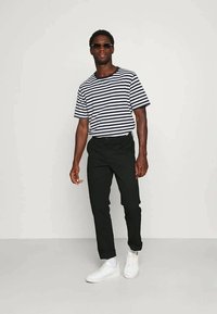 Striped short-sleeve t-shirt in navy and white, paired with black trousers. Model wears sunglasses and white sneakers, posing against a plain background.