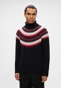 Homme portant un pull à col roulé noir avec un motif nordique rouge, blanc et gris, se tenant devant un fond clair uni.