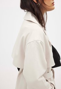 Šviesiai bežinė trenčkotų su struktūrizuota apykakle, kontrastinga nugaros panelė ir diržo detalė; glotnus paviršius ir laisvas siluetas.