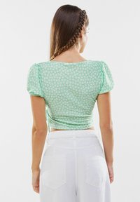 Top corto floreale verde con maniche a sbuffo, realizzato in un tessuto leggero, con motivi di fiori bianchi. Abbinato a pantaloni bianchi a vita alta.