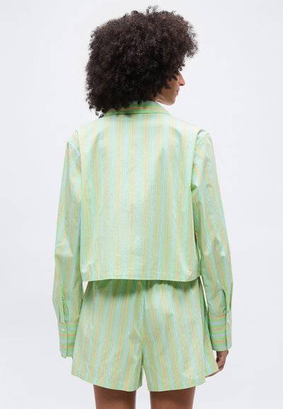 Άτομο με κατσαρά μαλλιά που φοράει μια μακρυμάνικη μπλούζα με κάθετες ρίγες σε πράσινο, μπλε και πορτοκαλί χρώμα και ταιριαστά σορτς, εμφανιζόμενο από πίσω.