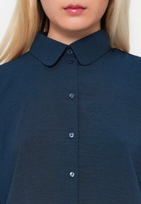 Cămașă bleu marin cu guler, având închidere cu nasturi și un material texturat. Gulerul are margini rotunjite.