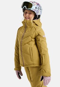 Gelbe isolierte Skijacke mit hohem Kragen, schrägen Nähten und zwei Seiten Taschen, kombiniert mit pinken Handschuhen und einem gemusterten Helm.