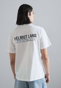 Valge puuvillane t-särk lühikeste varrukatega. Seljal on must tekst, sealhulgas brändi nimi "HELMut LANG" ja lisainformatsioon.
