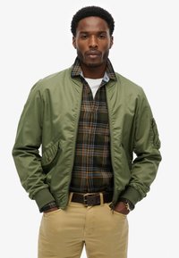 Chaqueta bomber verde oliva con bolsillos laterales, llevada sobre una camisa de cuadros oscura con acentos en amarillo y naranja, combinada con pantalones caqui.