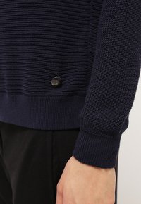 Pull en maille bleu marine avec rayures texturées, poignets et ourlet côtelés, comprenant un détail de bouton rond noir sur le côté.