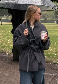 Mørkegrå indpakningsfrakke med brede ærmer og bælte, samt let blå jeans. Holder en kaffekop under en klar paraply.