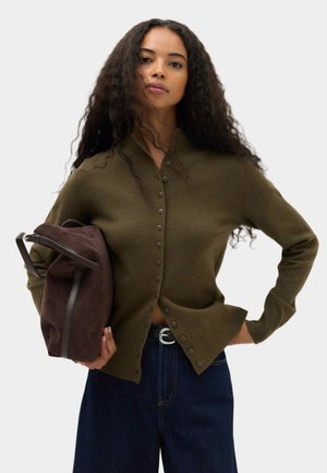 Frau mit lockigem Haar, die eine olivgrüne, geknöpfte Strickjacke und dunkle Jeans trägt und eine dunkelbraune Wildledertasche unter dem Arm hält.