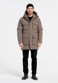 Brun, vadderad parka med huva, utrustad med fyra framfickor, tryckknappar och dragkedja. Buren över en svart tröja och byxor.