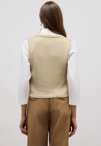 Top sin mangas de punto en brillo dorado, con un escote redondo y un diseño ajustado, combinado con una camisa blanca de mangas largas y pantalones marrones.