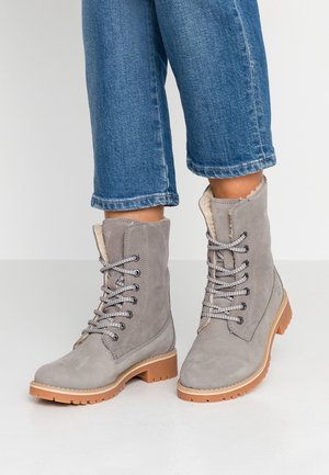 Bottes à lacets en daim gris clair avec doublure en fourrure et semelles épaisses couleur beige porté avec un jean bleu raccourci sur un sol blanc.