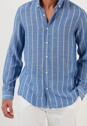 Man met een blauw overhemd met lange mouwen en witte verticale strepen, en een witte broek, hand gedeeltelijk zichtbaar met een tattoo.