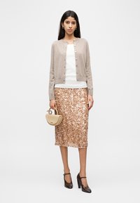 Donna che indossa un cardigan beige, una maglia bianca, una gonna midi con paillettes color oro rosa, tiene una borsa beige e indossa scarpe con tacco marroni, in piedi su sfondo bianco.
