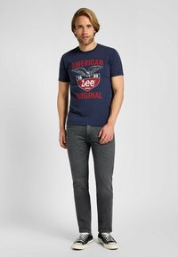 Camiseta de algodón azul marino con una impresión gráfica que presenta un águila y el texto "AMERICAN ORIGINAL LEE BRAND" en rojo, combinada con jeans grises y zapatillas negras.