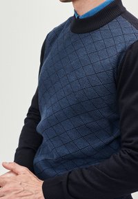 Uomo che indossa un maglione blu navy con un motivo a scacchiera testurizzato sopra una camicia blu con colletto, mani unite davanti.