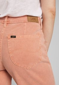 Jeans de mezclilla rosa con un bolsillo trasero que presenta un detalle de costura curva. Etiqueta de cuero de la marca adjunta a la cintura. Tejido texturizado.