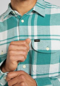 Chemise en flanelle à carreaux bleu sarcelle et blanc avec fermeture à boutons, dotée d'une poche poitrine et d'une petite étiquette noire portant le texte "Lee".