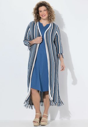 Donna sorridente con capelli ricci, che indossa un vestito blu al ginocchio e un lungo cardigan a righe con frange, in piedi davanti a uno sfondo neutro.