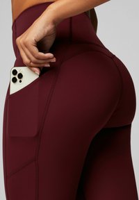 Burgunderfarbene Leggings aus dehnbarem Material, mit einer Seitentasche für ein Smartphone und sichtbaren Nähdetails.