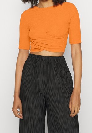 Vrouw die een oranje cropped top met korte mouwen en een twist detail draagt, gecombineerd met zwart, hoog getailleerd, geplooid, broek met wijde pijpen.