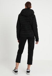 Donna con i capelli castani raccolti in un bun, che indossa una giacca a vento nera con cappuccio, pantaloni neri cropped e sneakers bianche Adidas, in piedi di spalle.