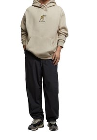 Beżowa bluza z kapturem z przednią kieszenią i żółtym detalem graficznym, zestawiona z luźnymi czarnymi spodniami. Obuwie to mieszanka czarnych i beżowych sneakersów.